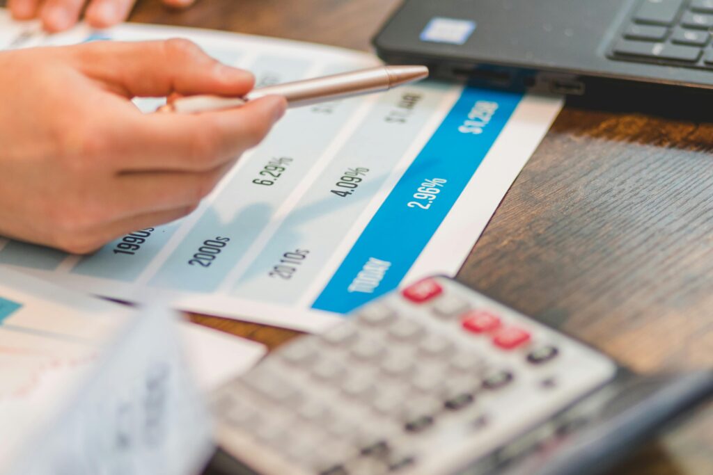 A financial report with a calculator and printed papers, a laptop computer and a hand pointing to profit loss with a pencil.