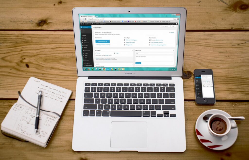 Laptop displaying a WordPress dashboard on a wooden desk, with a smartphone, notebook, pen, and cup of coffee beside it.